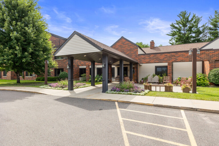 Benedict Village Entrance: A brick building with a covered entryway, surrounded by greenery and a paved path under clear skies.
