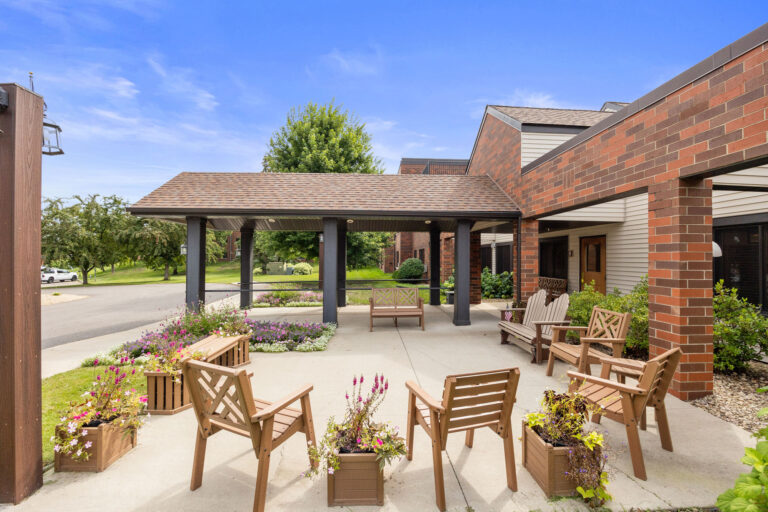 Outdoor patio with wooden chairs, a bench, and flower pots under a covered structure in front of the Benedict Village entrance.