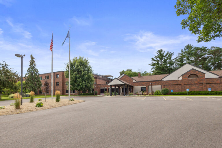 A brick building with a sloped roof marks the Benedict Village Entrance, surrounded by flags, a parking lot, and trees.