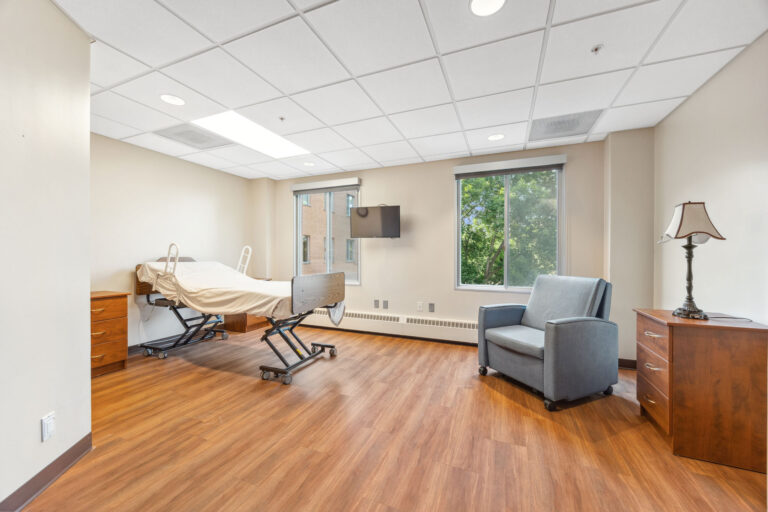 Long term care apartment with a bed, nightstands, armchair, TV, and a window showcasing treetops outside.