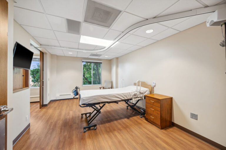 Long term care apartment with a bed, nightstands, armchair, TV, and a window showcasing treetops outside.