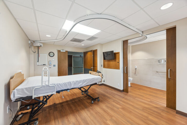 Long-term care apartment with an adjustable bed, wooden floors, wall-mounted TV, and adjacent bathroom.