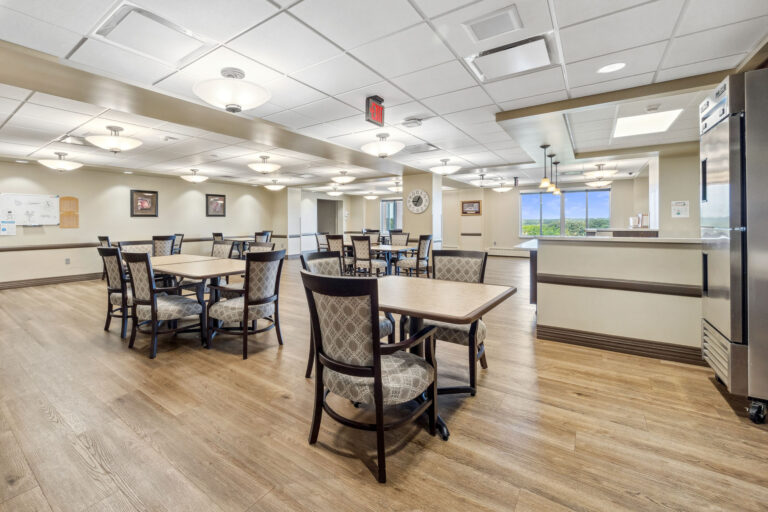 Spacious long-term care dining room with wooden floors, square tables, cushioned chairs, and bright overhead lighting.