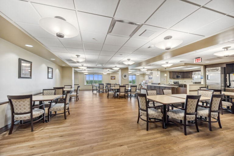 Spacious long-term care dining room with wooden floors, square tables, cushioned chairs, and bright overhead lighting.