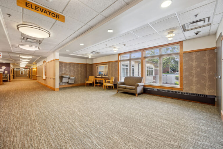 Long term care facility lounge area includes pacious hallway with chairs, a table, and elevator sign,