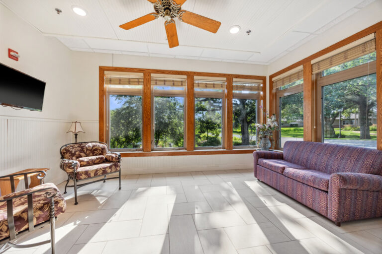 The long term care sunroom is a sunlit living room with large windows, a sofa, and ceiling fan, overlooking a green lawn and trees.
