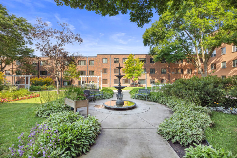 Lillians Garden: A courtyard with a central fountain, lush greenery, benches, and a brick building in the background.