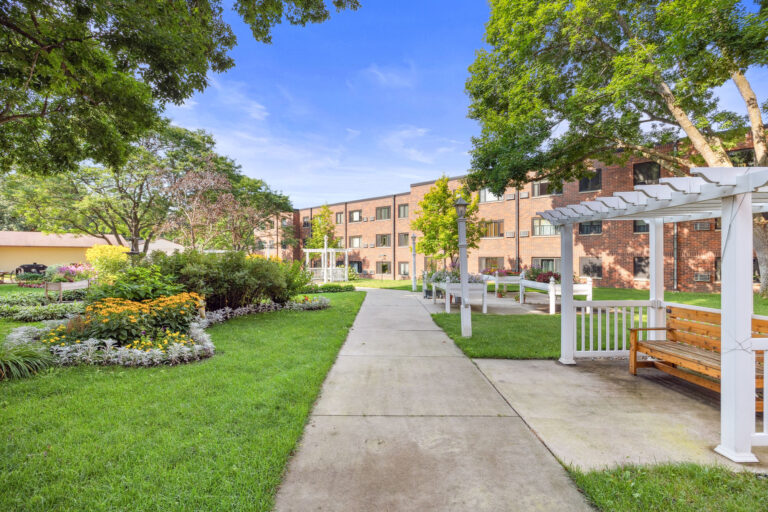 Lillians Garden features a walking path with benches and pergolas, leading to a brick building under the blue sky.