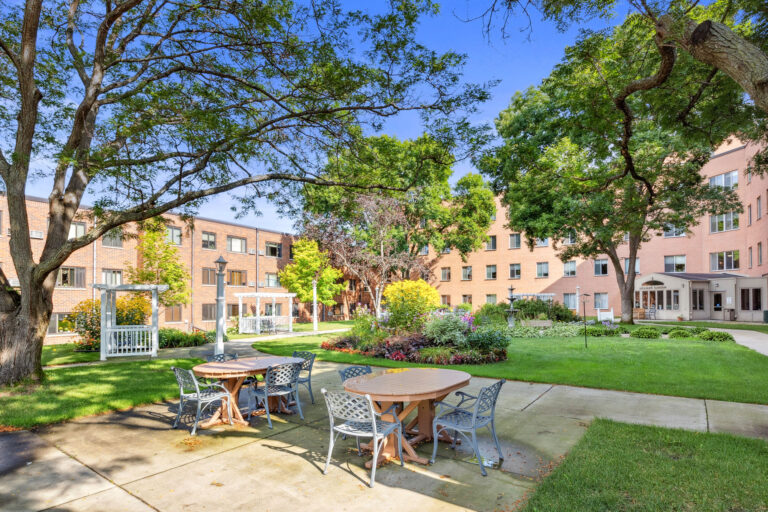Lillians Garden Patio: A courtyard with tables, chairs, trees, and a brick building under a clear blue sky.