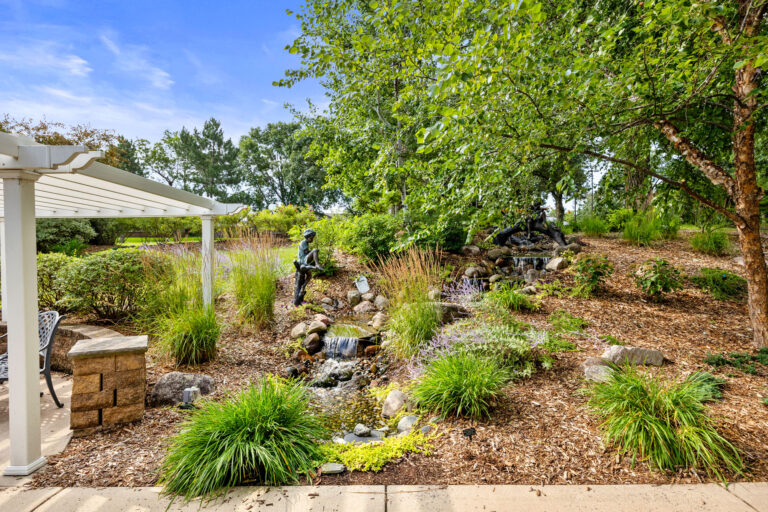 Rosies Garden boasts a pergola, lush greenery, and statues around a charming water feature under a clear blue sky.