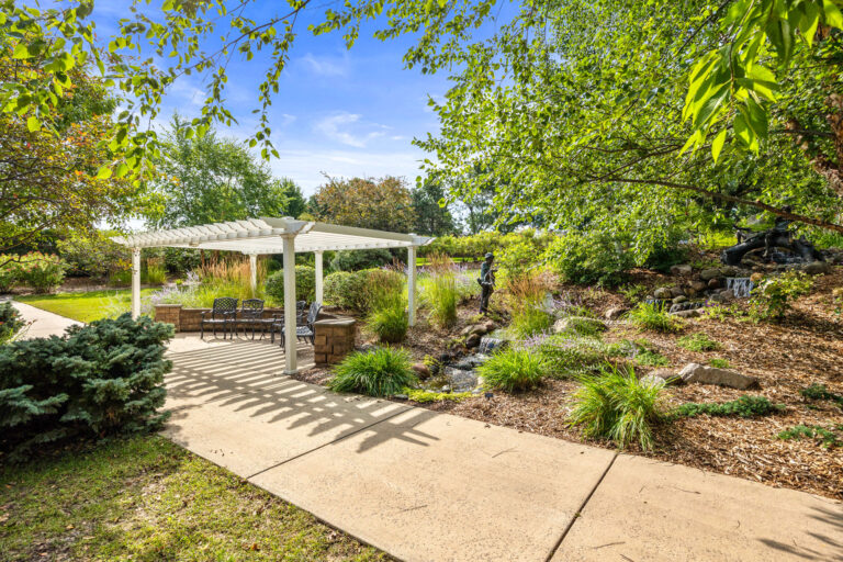 Rosies Garden Seating Area: A pergola over patio furniture, surrounded by lush greenery and a small pond with statues.