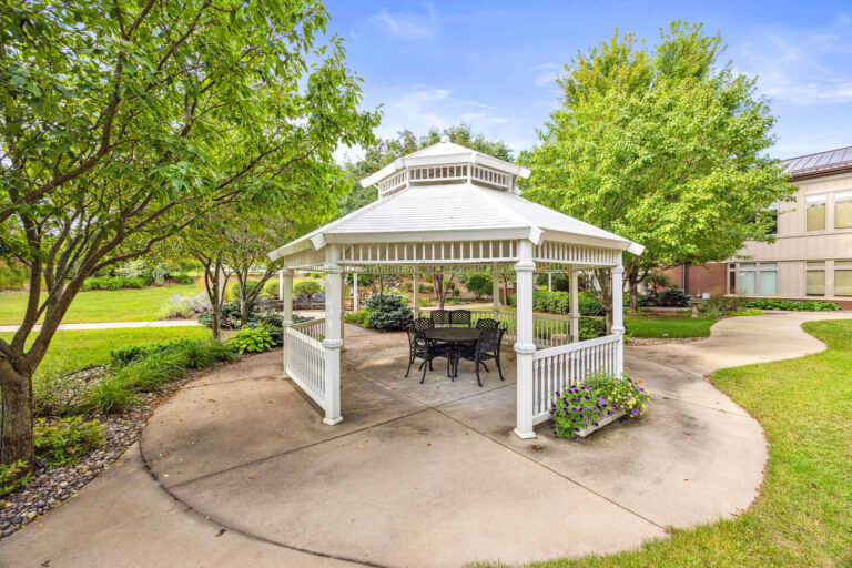Rosies Garden Gazebo hosts a cozy table and chairs in a lush setting, embraced by trees with a building in the background.