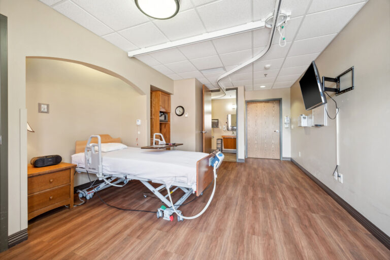 Short-term care apartment with an adjustable bed, wooden floors, wall-mounted TV, and adjacent bathroom.