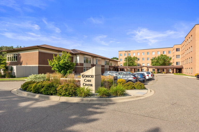 Exterior of Gorecki Transitional Care Center entrance with a parking lot and cars on a sunny day.