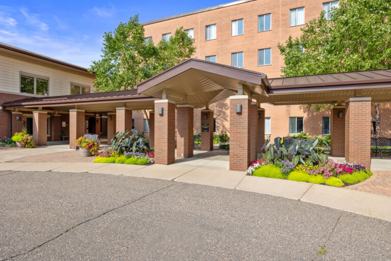 Transitional Care Center entrance with covered walkway, surrounded by colorful plants and trees on a sunny day.