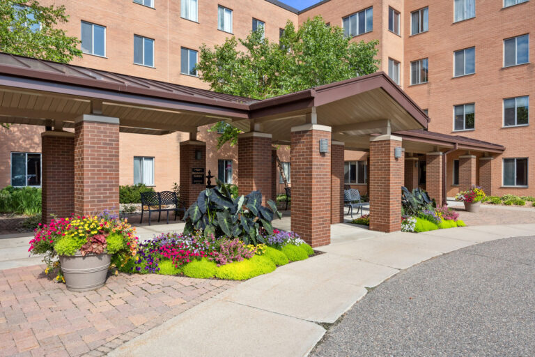 Transitional Care Center entrance with covered walkway, surrounded by colorful plants and trees on a sunny day.