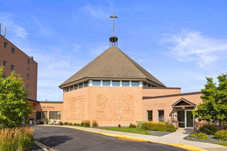 St. Benedict's main entrance is a brick church with a conical roof and cross, surrounded by greenery under a blue sky.
