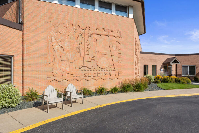 St. Benedicts main entrance features a brick building with an engraved mural and two white chairs out front.