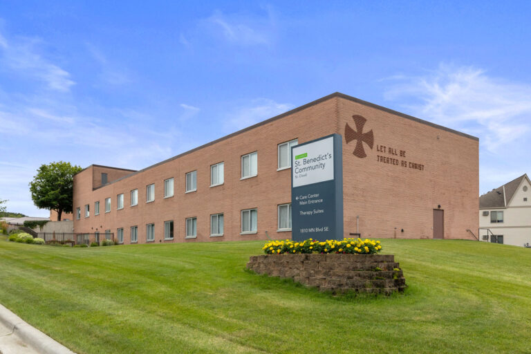 Brick building of St. Benedicts Community with flowers in front, marking the Main Entrance on a grassy hill.