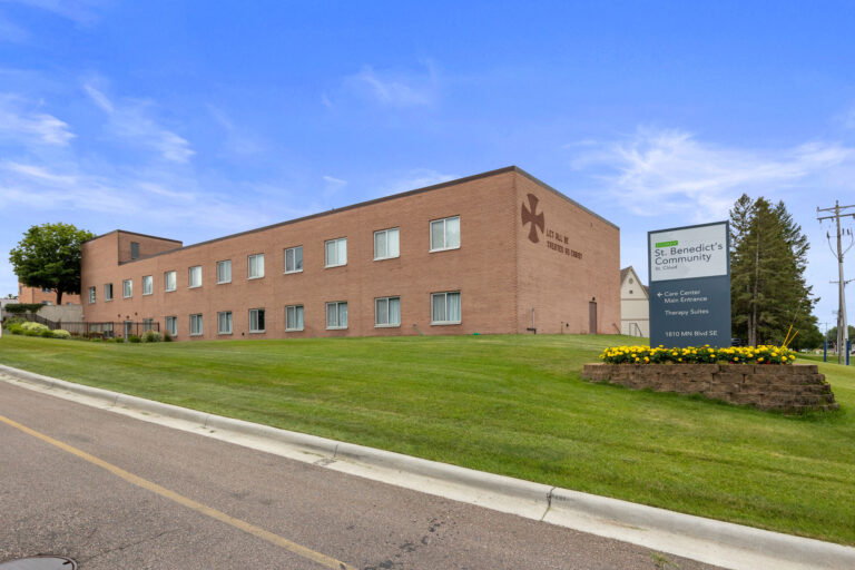Brick building of St. Benedicts Community with flowers in front, marking the Main Entrance on a grassy hill.