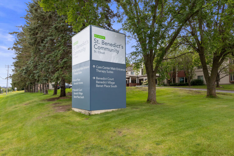 Monument sign with Ecumen St. Benedict's Community logo with directional arrows to specific community driveways.